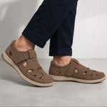 A pair of taupe-brown cutout sandals with hook-and-loop straps, shown on a model wearing dark navy pants against a neutral gray studio background.