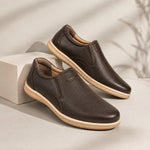 A pair of dark brown slip-on leather shoes with beige soles, displayed on a light gray surface with a white geometric block and soft shadows.