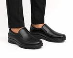 A pair of men's black slip-on leather shoes worn with black trousers, shown from a side angle against a clean white background.