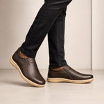 A person wearing dark brown leather slip-on loafers with tan soles, paired with black jeans, shown from the knees down against a neutral beige studio background.