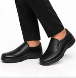 A pair of black slip-on work shoes worn with black trousers, shown from a side profile against a clean white background.